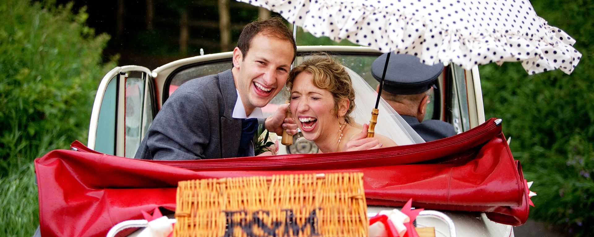 Newlyweds laughing in the rain with umbrellas, sitting in the back of Cassie with the roof down